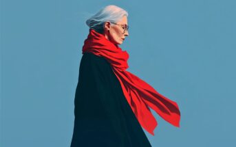 An older woman with white hair and glasses, wearing a flowing red scarf and a dark coat, stands in profile against a plain blue background. The scarf appears to be blowing in the wind.