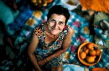 A smiling woman in a patterned dress sits on a colorful picnic blanket, surrounded by baskets of fruit, looking up at the camera. The atmosphere is bright and cheerful.