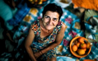 A smiling woman in a patterned dress sits on a colorful picnic blanket, surrounded by baskets of fruit, looking up at the camera. The atmosphere is bright and cheerful.
