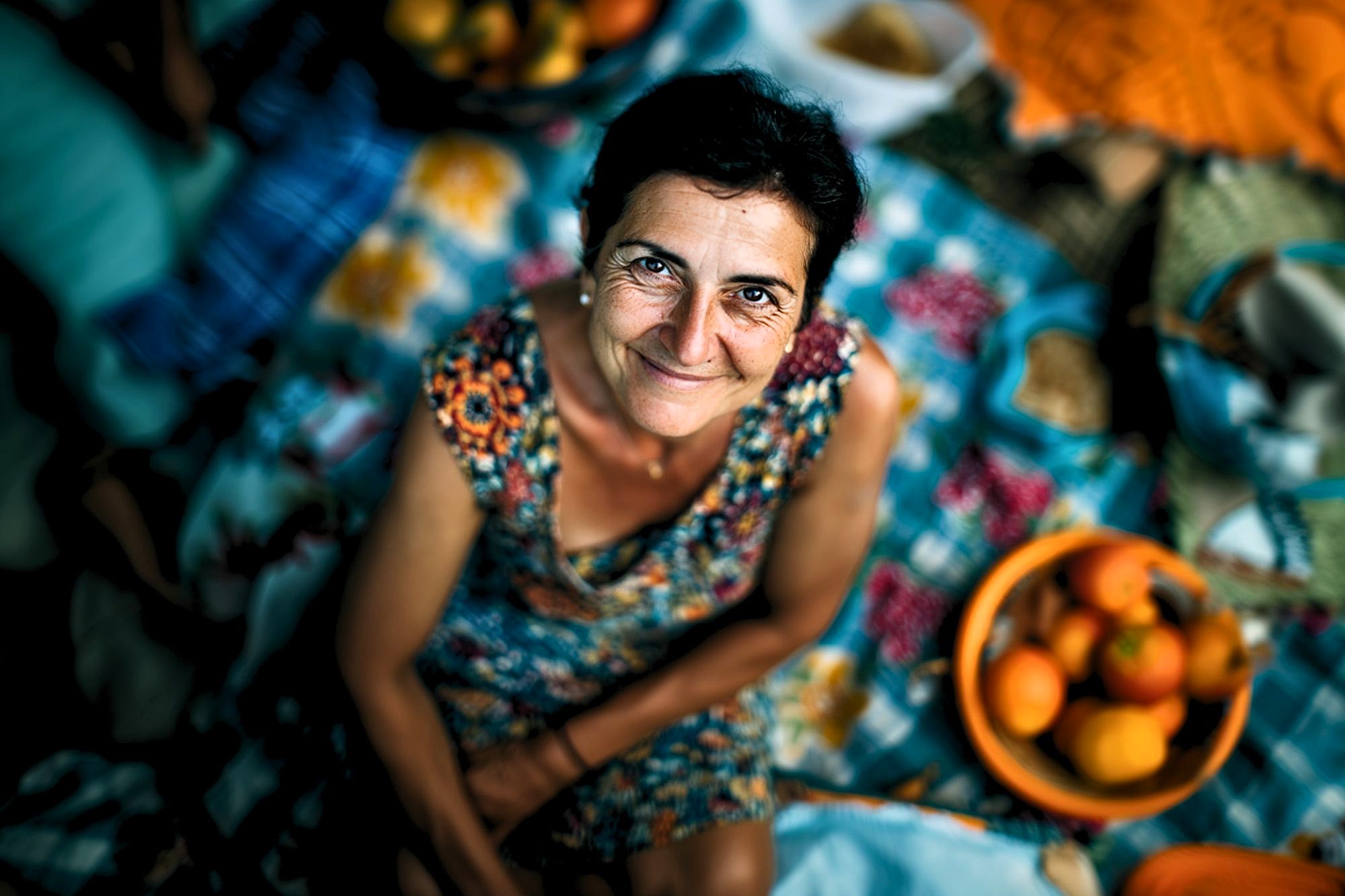 A smiling woman in a patterned dress sits on a colorful picnic blanket, surrounded by baskets of fruit, looking up at the camera. The atmosphere is bright and cheerful.