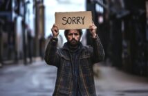 A man in a plaid jacket stands in an empty street, holding up a cardboard sign that reads "SORRY" in large letters. He has a serious expression and appears to be seeking forgiveness.