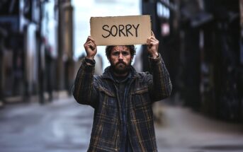 A man in a plaid jacket stands in an empty street, holding up a cardboard sign that reads "SORRY" in large letters. He has a serious expression and appears to be seeking forgiveness.