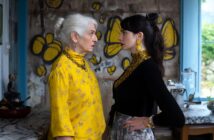 An older woman in a yellow outfit and a younger woman in black and yellow stand face-to-face in a kitchen, staring intently at each other. Yellow butterfly graffiti decorates the wall behind them.