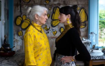 An older woman in a yellow outfit and a younger woman in black and yellow stand face-to-face in a kitchen, staring intently at each other. Yellow butterfly graffiti decorates the wall behind them.