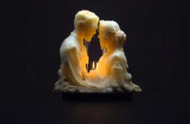 A candle sculpture of a man and woman facing each other, their faces melting from the flame burning between them, creating a warm, dramatic glow in the dark.