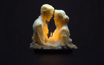 A candle sculpture of a man and woman facing each other, their faces melting from the flame burning between them, creating a warm, dramatic glow in the dark.