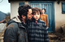 A man gently kisses a boy on the head, standing close together outside a rustic house. The boy looks directly at the camera, while a woman stands blurred in the background near a blue door.