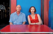 A man in a blue shirt and a woman in a red dress sit side by side at a red table in front of a bright blue and red wall, both looking directly at the camera with serious expressions.