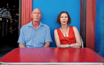 A man in a blue shirt and a woman in a red dress sit side by side at a red table in front of a bright blue and red wall, both looking directly at the camera with serious expressions.