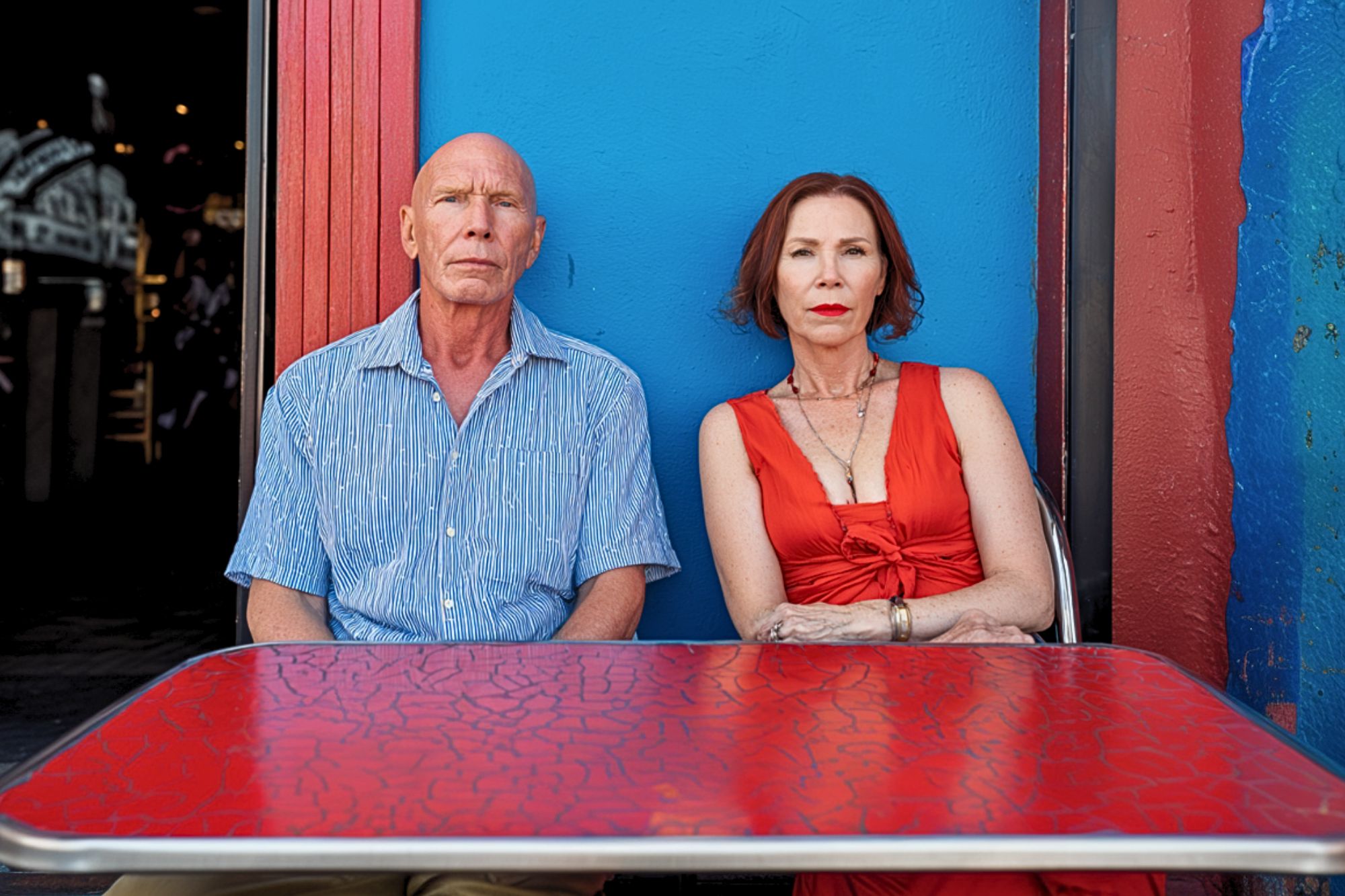 A man in a blue shirt and a woman in a red dress sit side by side at a red table in front of a bright blue and red wall, both looking directly at the camera with serious expressions.