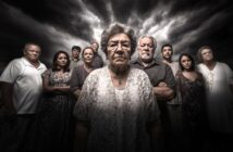 A group of serious-looking people, mostly older adults, stand closely together under a dramatic, cloudy sky. The woman in front wears glasses and a patterned dress, appearing somber and resolute.