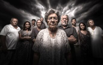 A group of serious-looking people, mostly older adults, stand closely together under a dramatic, cloudy sky. The woman in front wears glasses and a patterned dress, appearing somber and resolute.