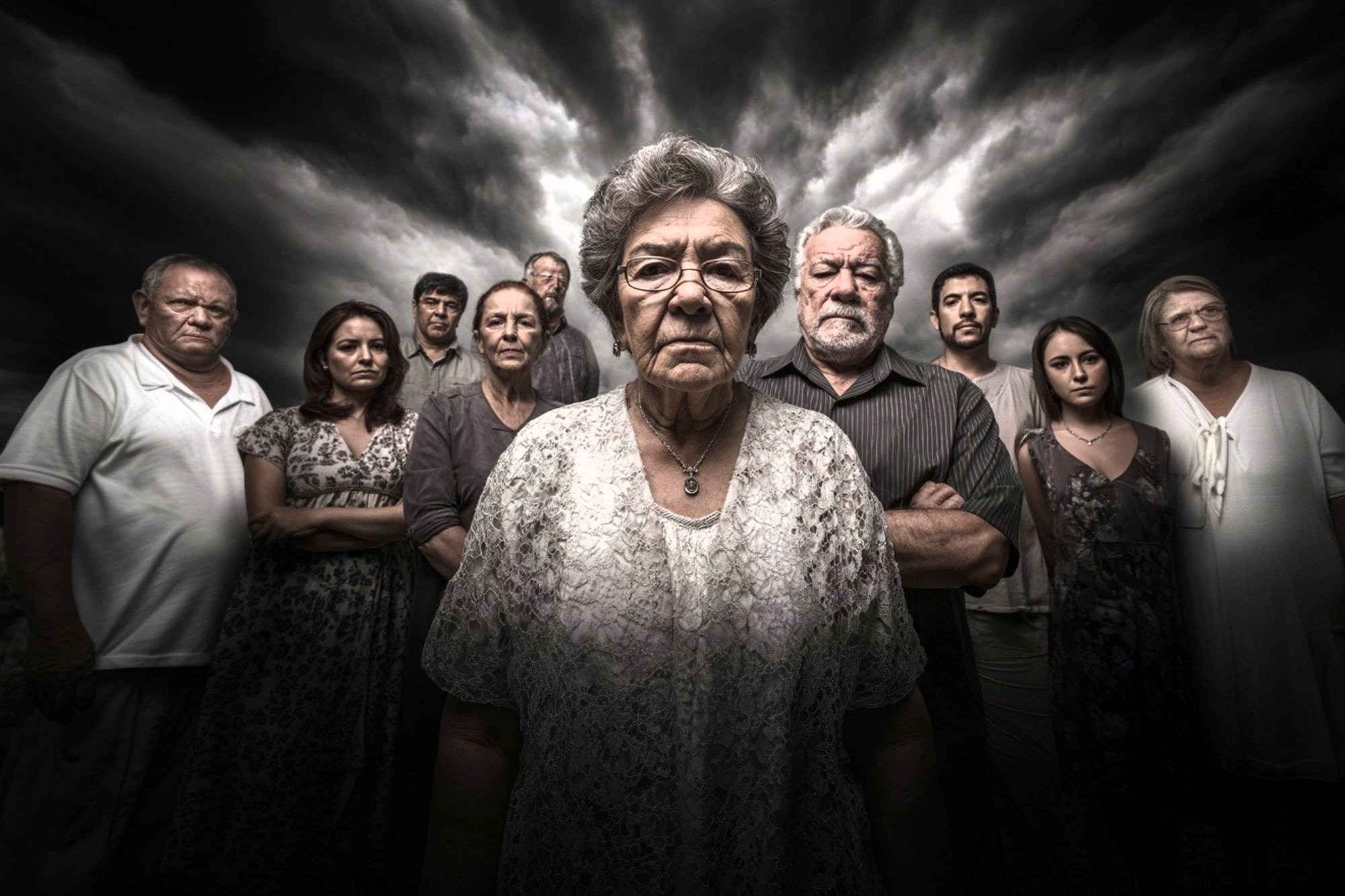A group of serious-looking people, mostly older adults, stand closely together under a dramatic, cloudy sky. The woman in front wears glasses and a patterned dress, appearing somber and resolute.