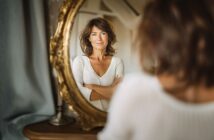 A woman with brown hair and a white sweater stands with arms crossed, looking confidently at her reflection in an ornate gold-framed mirror. The setting appears to be a cozy, softly lit room.