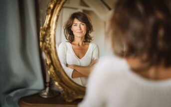A woman with brown hair and a white sweater stands with arms crossed, looking confidently at her reflection in an ornate gold-framed mirror. The setting appears to be a cozy, softly lit room.
