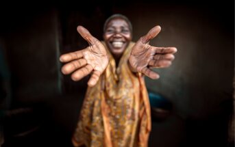A smiling person in patterned clothing extends their open hands toward the camera, palms facing forward. The background is dark and blurred, highlighting the joyful expression and welcoming gesture.