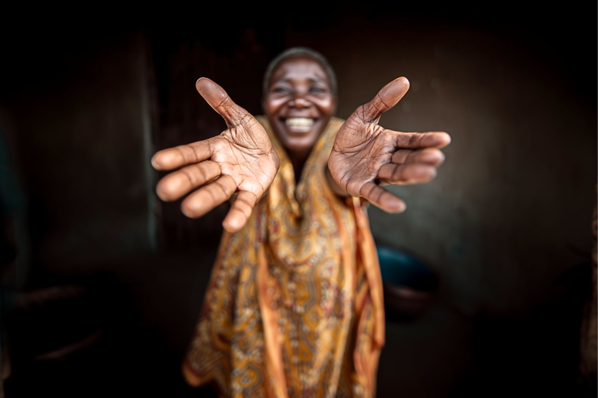 A smiling person in patterned clothing extends their open hands toward the camera, palms facing forward. The background is dark and blurred, highlighting the joyful expression and welcoming gesture.