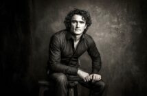 A man with curly hair wearing a dark shirt and jeans sits on a stool against a textured, dark background, looking seriously at the camera. The image is black and white and has a dramatic, moody lighting.