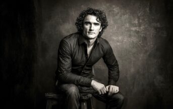 A man with curly hair wearing a dark shirt and jeans sits on a stool against a textured, dark background, looking seriously at the camera. The image is black and white and has a dramatic, moody lighting.