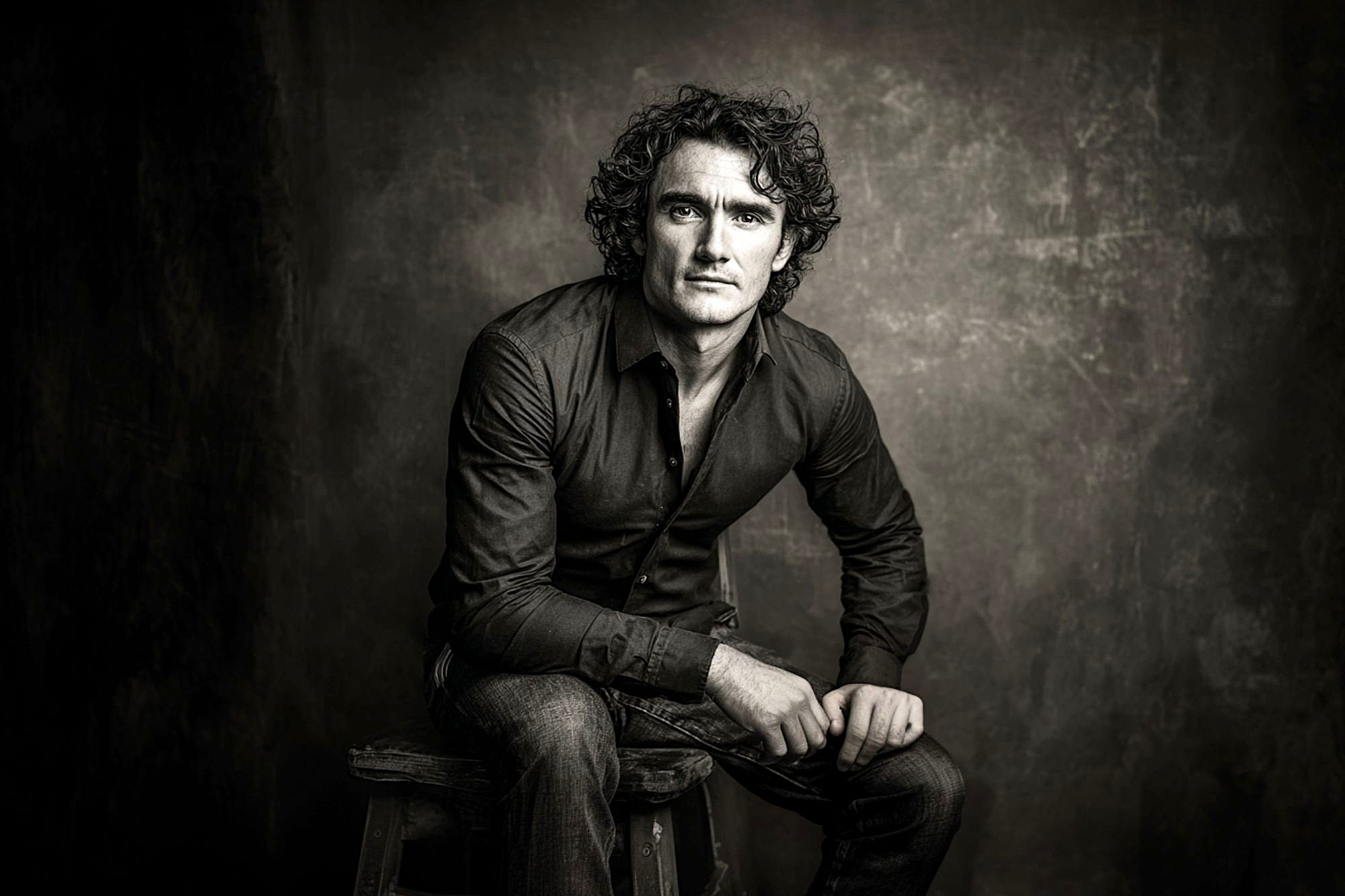 A man with curly hair wearing a dark shirt and jeans sits on a stool against a textured, dark background, looking seriously at the camera. The image is black and white and has a dramatic, moody lighting.