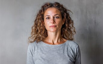 A woman with curly brown hair wearing a light gray long-sleeve shirt stands against a plain gray background, looking directly at the camera with a neutral expression.
