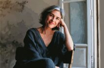 A woman with short gray hair and a black outfit sits by a window, smiling gently with her head resting on her hand. The lighting is soft and the scene has a painterly effect.
