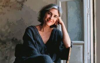 A woman with short gray hair and a black outfit sits by a window, smiling gently with her head resting on her hand. The lighting is soft and the scene has a painterly effect.