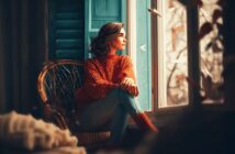A woman in a red sweater sits in a wicker chair by a window, gazing outside. Soft light streams in, and white paper birds appear to be flying past, creating a dreamy, tranquil atmosphere.