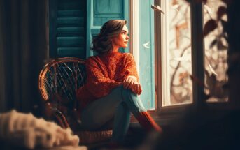 A woman in a red sweater sits in a wicker chair by a window, gazing outside. Soft light streams in, and white paper birds appear to be flying past, creating a dreamy, tranquil atmosphere.