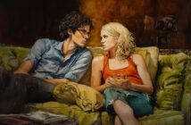 A man and woman sit close together on a green couch, gazing at each other intently. The woman holds a glass, and both appear engaged in a serious conversation in a warmly lit room.