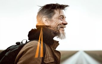 A bearded man with a backpack smiles in profile outdoors; a graphic of a road leads into the horizon, blended into his upper body and background, symbolizing a journey or adventure.