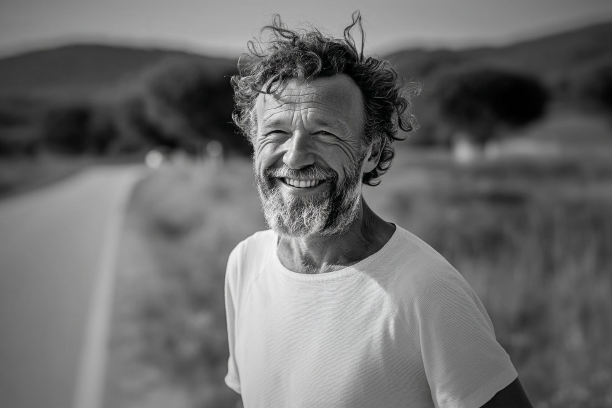 A smiling older man with a beard stands by a rural road, wearing a plain white t-shirt. The background is blurred, showing trees and hills on a sunny day. The photo is in black and white.