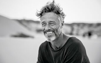 A middle-aged man with wavy hair and a beard smiles warmly while sitting outdoors on a sandy beach. The photo is black and white, creating a relaxed and natural atmosphere.