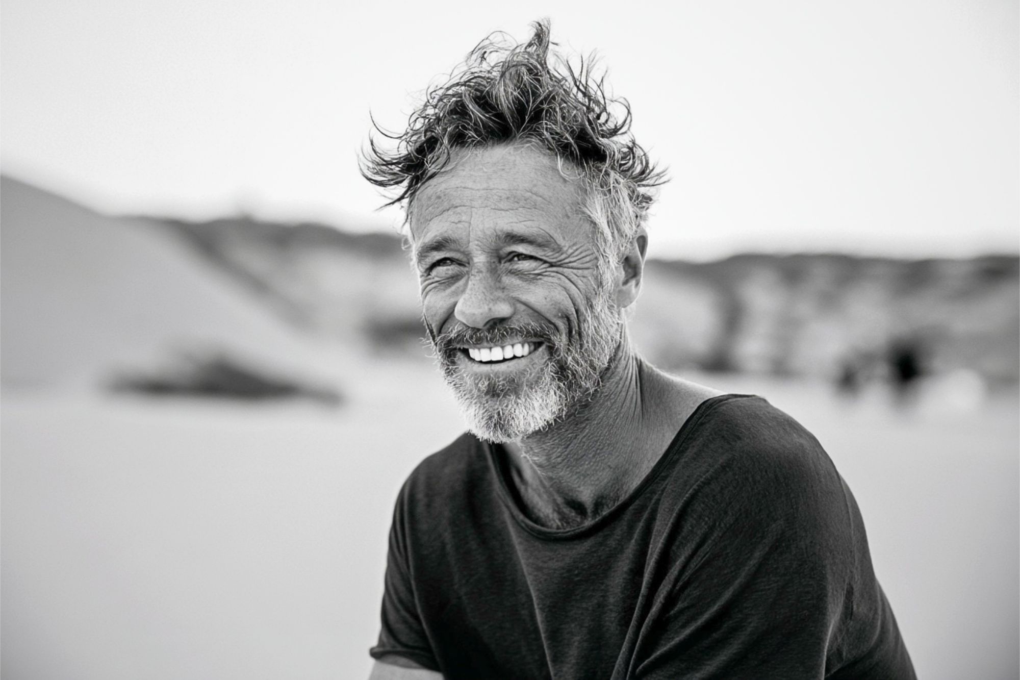 A middle-aged man with wavy hair and a beard smiles warmly while sitting outdoors on a sandy beach. The photo is black and white, creating a relaxed and natural atmosphere.