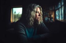 A woman with curly blonde hair sits indoors, gazing pensively out a window with soft natural light illuminating her face, creating a contemplative mood in a dimly lit room.