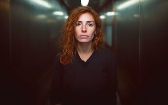 A woman with wavy red hair and freckles stands in a dimly lit hallway, looking directly at the camera with a neutral expression. She is wearing a dark, long-sleeved shirt and the background appears blurred.