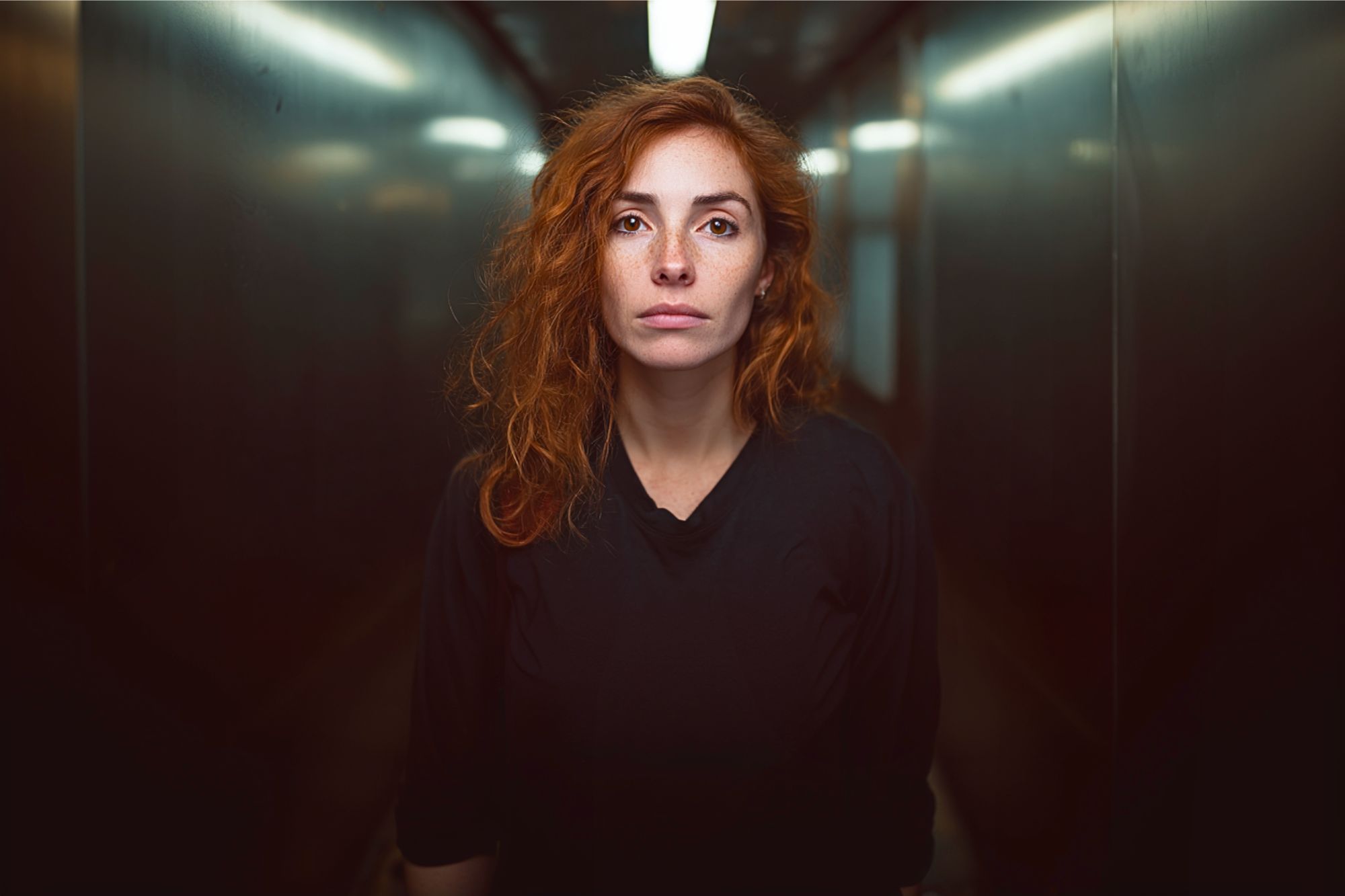 A woman with wavy red hair and freckles stands in a dimly lit hallway, looking directly at the camera with a neutral expression. She is wearing a dark, long-sleeved shirt and the background appears blurred.