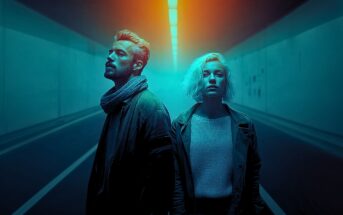 A man and a woman stand side by side in a dimly lit tunnel with blue and orange lighting, both looking serious and facing slightly away from each other. The tunnel is empty and atmospheric.