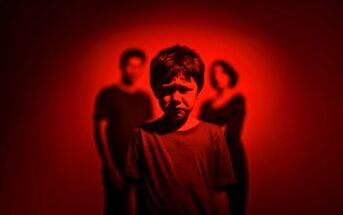 A young boy stands in the foreground with a sad expression, while two adults stand blurred in the background. The entire scene is illuminated with a dramatic red light.