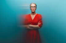 A confident older woman with gray hair in a bun and red glasses stands with arms crossed, wearing a red dress against a smooth blue background with a motion blur effect around her.