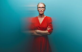 A confident older woman with gray hair in a bun and red glasses stands with arms crossed, wearing a red dress against a smooth blue background with a motion blur effect around her.