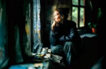 An elderly woman sits by a window with her chin resting on her hand, gazing outside thoughtfully. Soft light illuminates her face. A cup and some papers sit on the windowsill beside her.