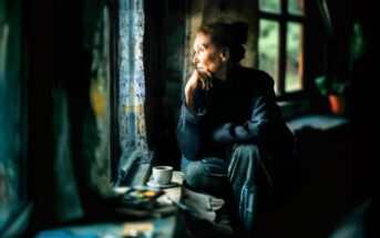 An elderly woman sits by a window with her chin resting on her hand, gazing outside thoughtfully. Soft light illuminates her face. A cup and some papers sit on the windowsill beside her.
