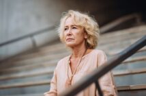 An older woman with blonde hair and a light beige blouse sits on outdoor steps, looking ahead with a thoughtful, serious expression. The background is blurred, focusing attention on her face.