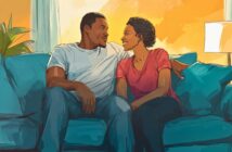 A couple sits close together on a blue couch, smiling and gazing at each other warmly in a cozy, sunlit living room with a lamp and plant in the background.