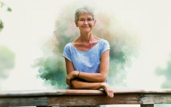 An older woman with short gray hair and glasses smiles while leaning on a wooden railing, wearing a light blue top with a blurred, pastel-colored background.