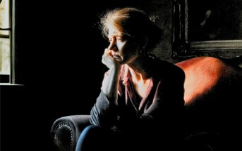 A woman sits alone on a chair in a dimly lit room, resting her head on her hand and gazing out a window, appearing deep in thought.