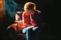 A person with light hair sits in a dimly lit room on a cushioned chair, leaning forward with their head resting on one hand, appearing deep in thought or sadness. Warm light highlights their hair and shirt.