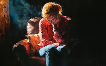 A person with light hair sits in a dimly lit room on a cushioned chair, leaning forward with their head resting on one hand, appearing deep in thought or sadness. Warm light highlights their hair and shirt.