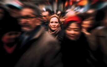 A woman stands still and looks ahead in a crowded, busy street. The people around her appear blurred, emphasizing her calm and focused presence amid the moving crowd.